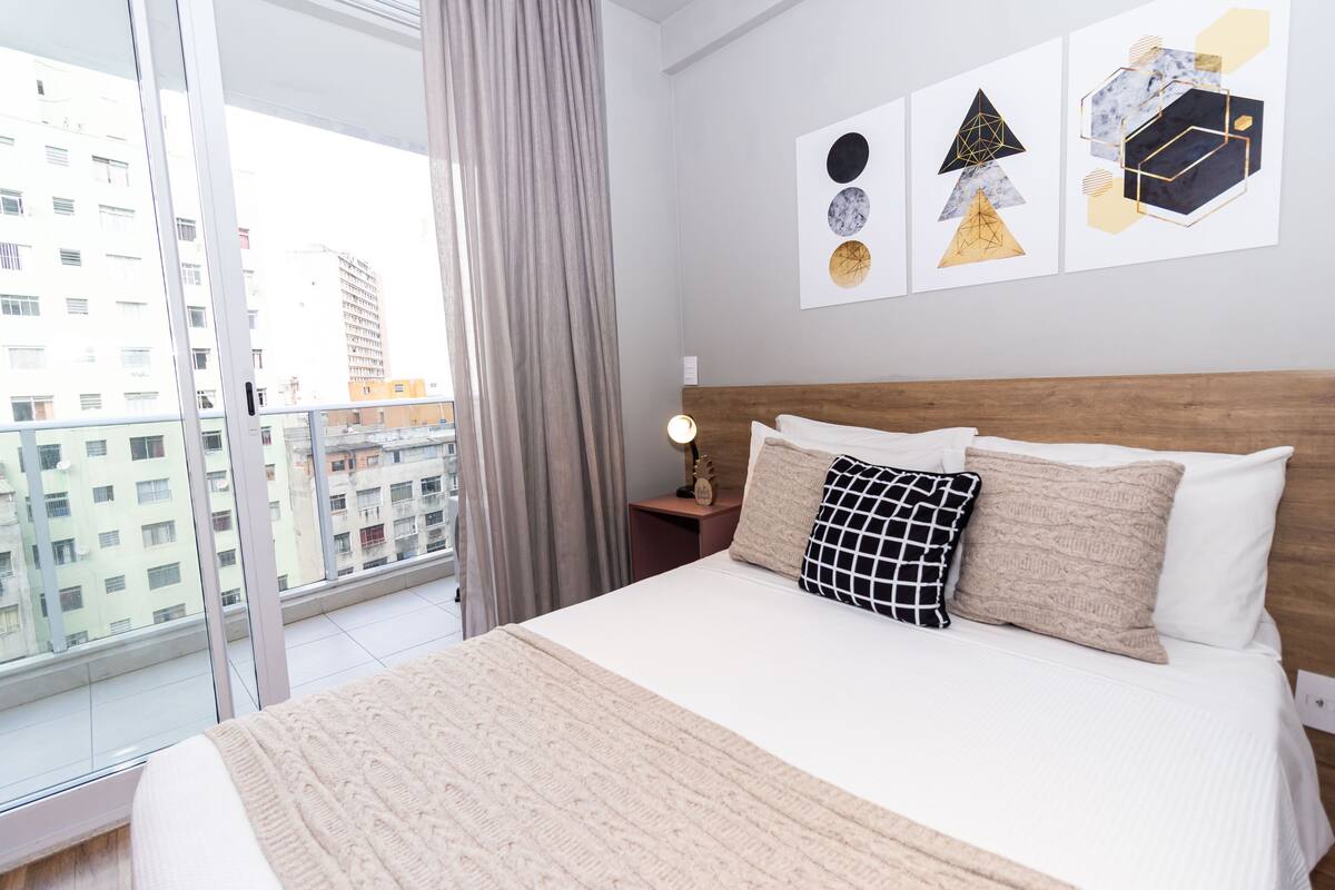 A bedroom with a large bed, a nightstand, a lamp, and a view of the city through a window.