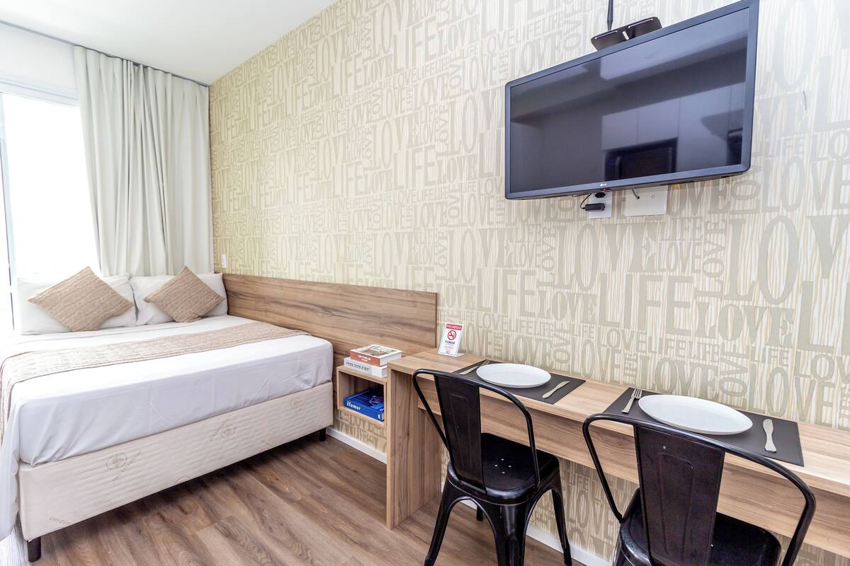 A hotel room with a bed, a desk with a chair, a TV mounted on the wall, and a wall with words like "love" and "life".