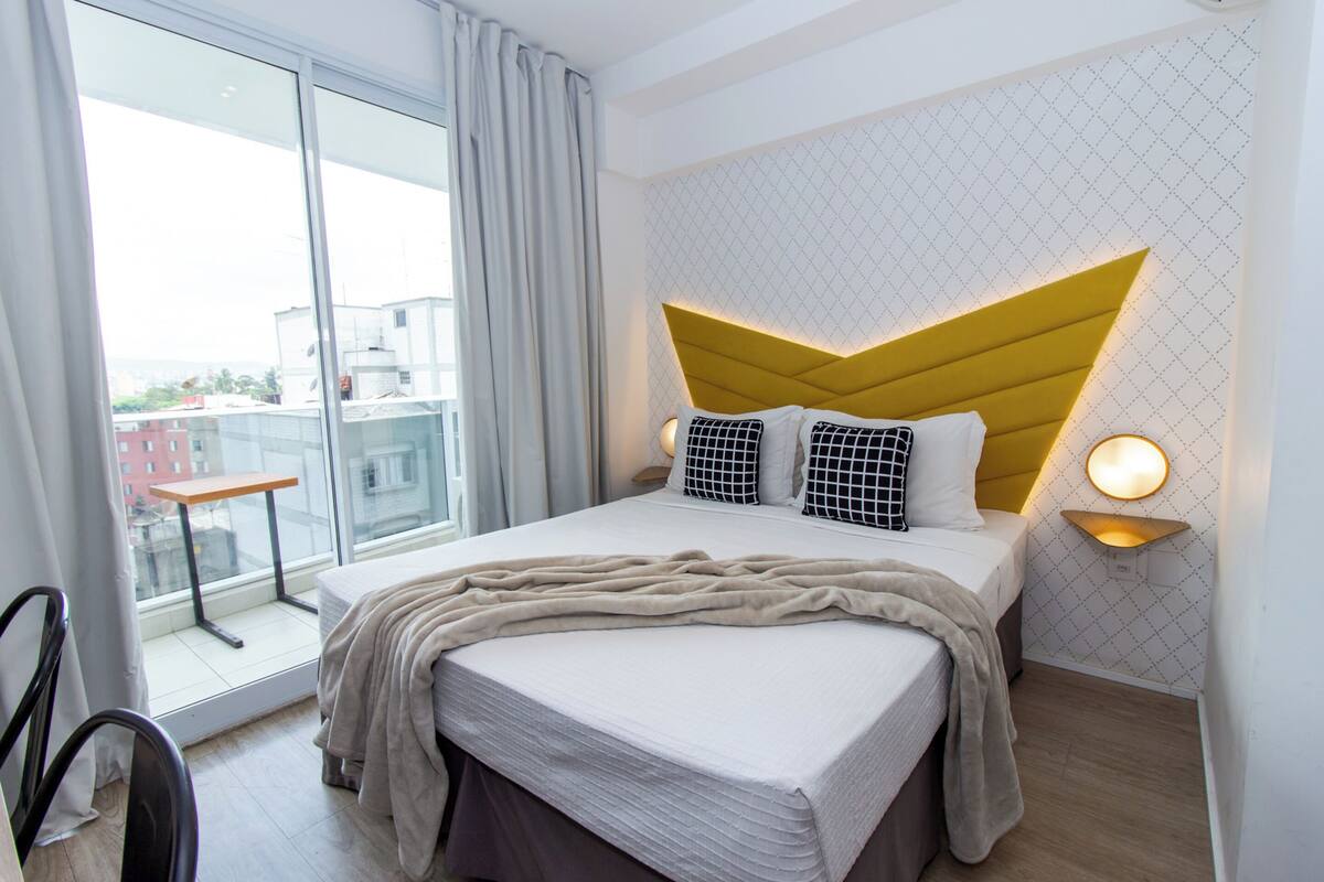 A modern hotel room with a large bed, a view of the city, a small table by the window, and a unique geometric headboard.