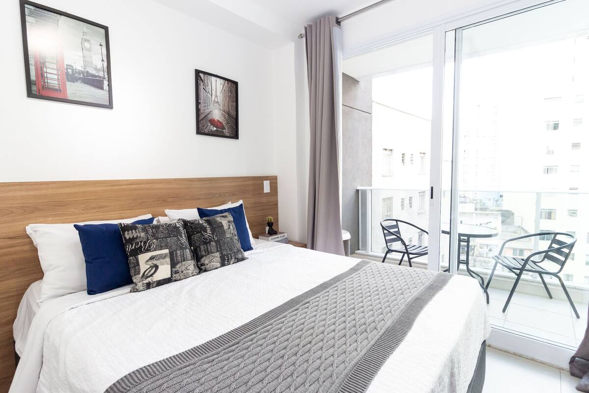 A hotel room with a large bed, a wooden headboard, two framed pictures on the wall, a balcony with chairs, and a view of buildings.