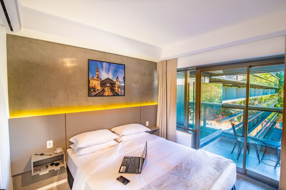 A modern hotel room with a large bed, a laptop, and a smartphone on the bed. There is a small bedside table with a cup and a pair of glasses. The room has a large window with a view of a balcony and outdoor seating.