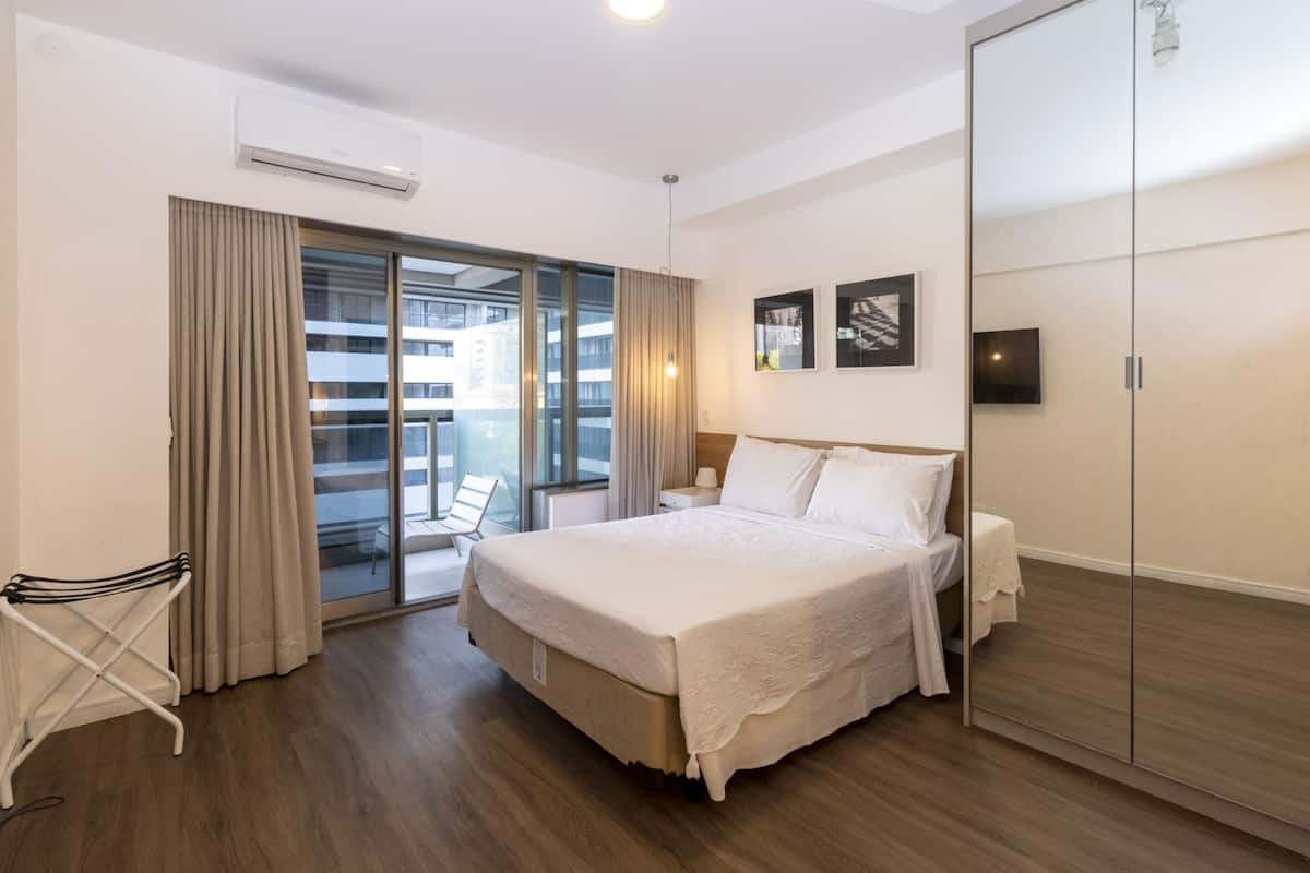 A hotel room with a large bed, a sliding glass door to a balcony, a white chair, and a wardrobe.