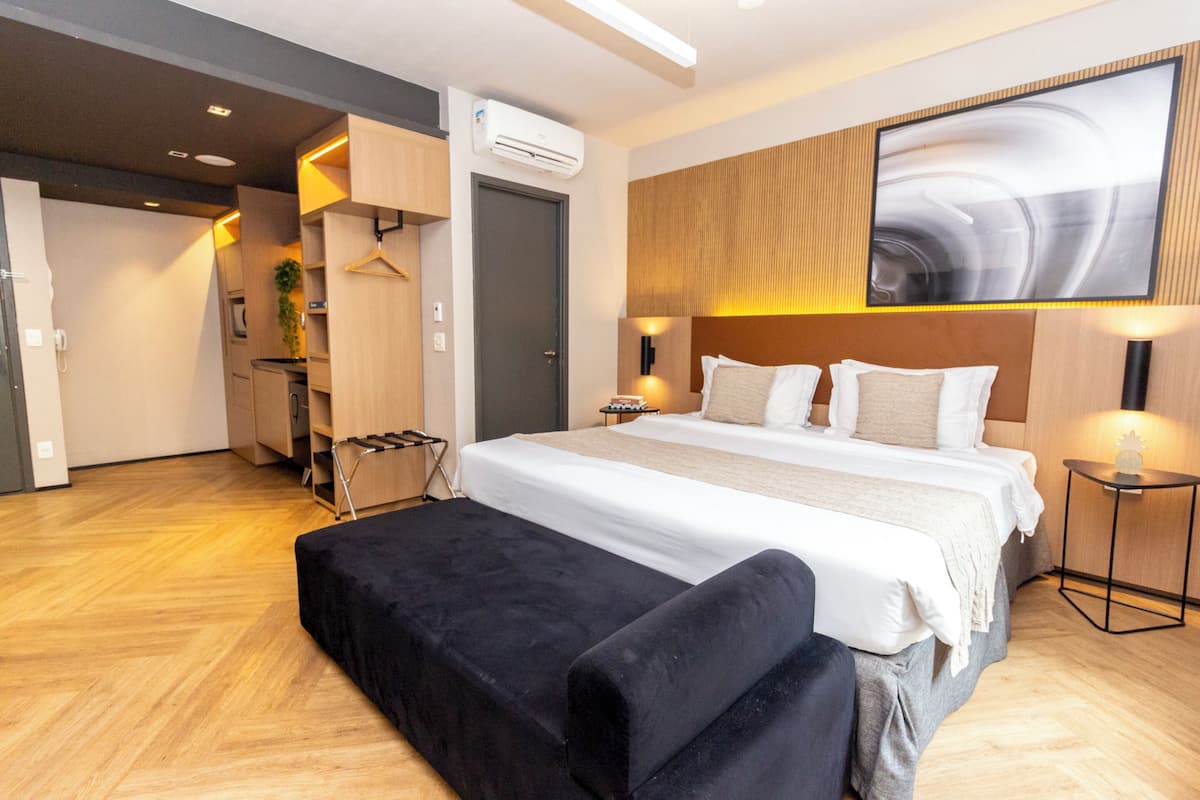 A modern hotel room with a large bed, a black ottoman, a wooden headboard, and a wall-mounted TV.