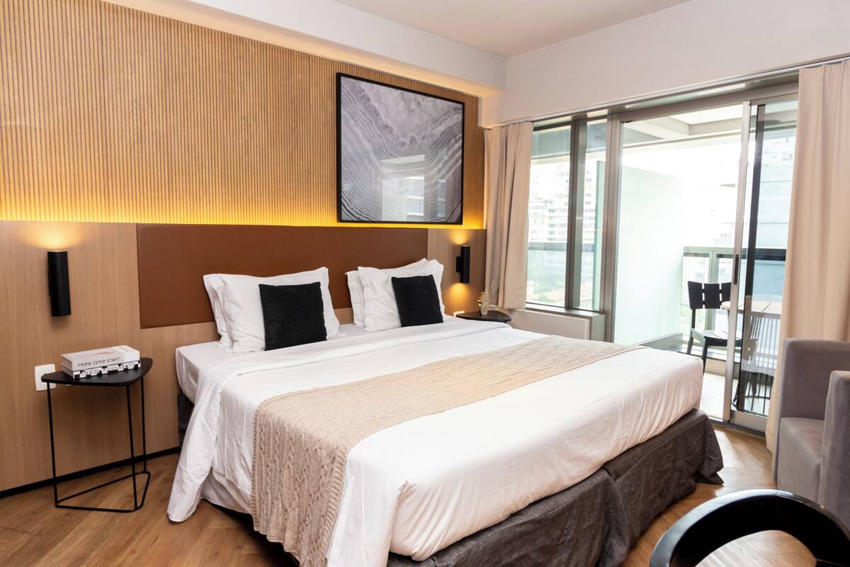 A hotel room with a large bed, a side table, a wall-mounted TV, and a balcony with a chair.