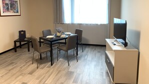 Ett modernt, väl upplyst rum med ett matbord, fyra stolar, en tv och ett litet skrivbord med en dator.