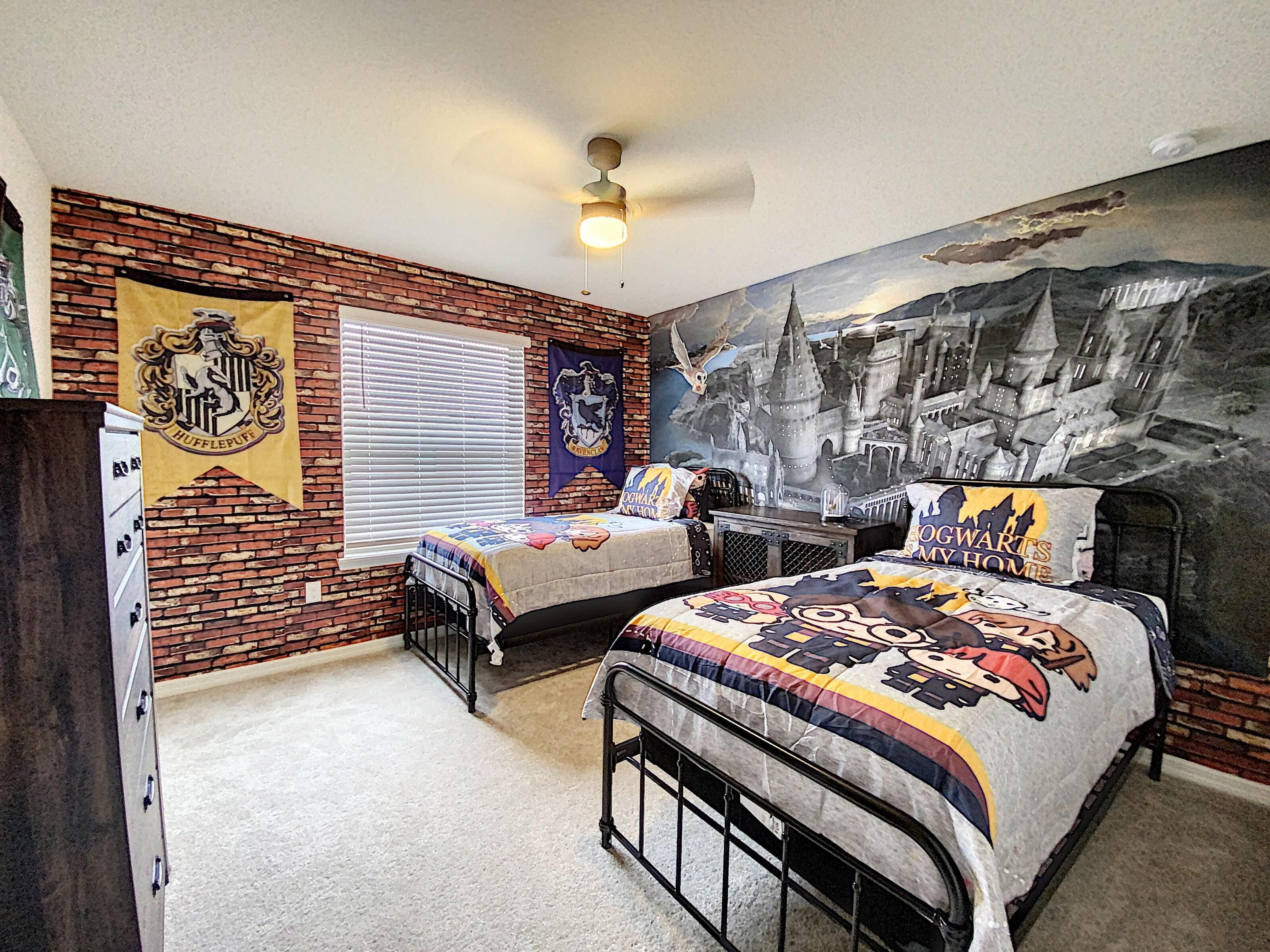 A bedroom with a brick wall, a ceiling fan, a bed with a Hogwarts-themed bedspread, and a wall mural of a castle.