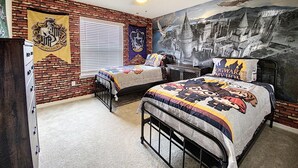 Una camera da letto con parete in mattoni, ventilatore a soffitto, un letto con copriletto a tema Hogwarts e un murale raffigurante un castello.