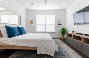 Quarto moderno com cama, uma janela grande com persianas, uma TV fixada na parede e uma pequena planta sobre uma mesinha de cabeceira.