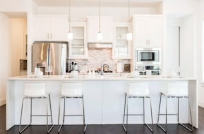 Uma cozinha moderna com armários brancos, uma ilha central com banquetas, eletrodomésticos de aço inoxidável e iluminação pendente.