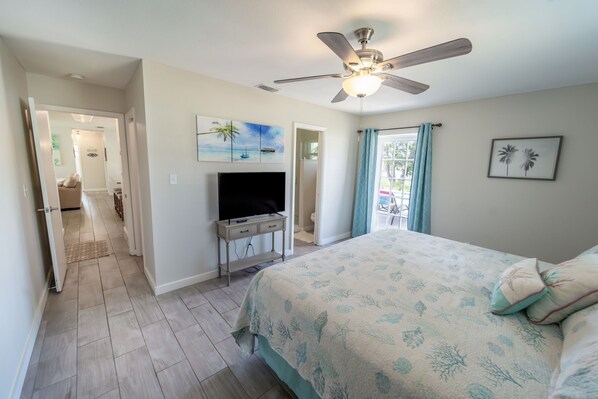 A bedroom with a bed, a ceiling fan, a TV on a stand, and a door leading to another room.