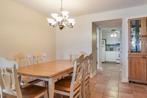 A dining area with a wooden table and white chairs, a chandelier, and a kitchen in the background.