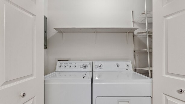 A laundry room with two stacked washers and dryers, a metal shelf above, and a white door with a lock. - Delightful Dolphin: Sound Views, Shared Pool & Tennis Court, Short Stays Anytime (Nags Head)