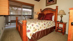 A bedroom with a wooden bed, a nightstand with a lamp, a window with blinds, and a framed picture on the wall. - Sierra Park Villas #82 (Mammoth Lakes)