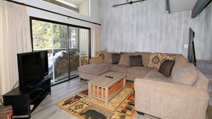 A living room with a sectional sofa, a flat-screen TV on a stand, a sliding glass door leading to a balcony, and a patterned rug. - Sierra Park Villas #93 (Mammoth Lakes)