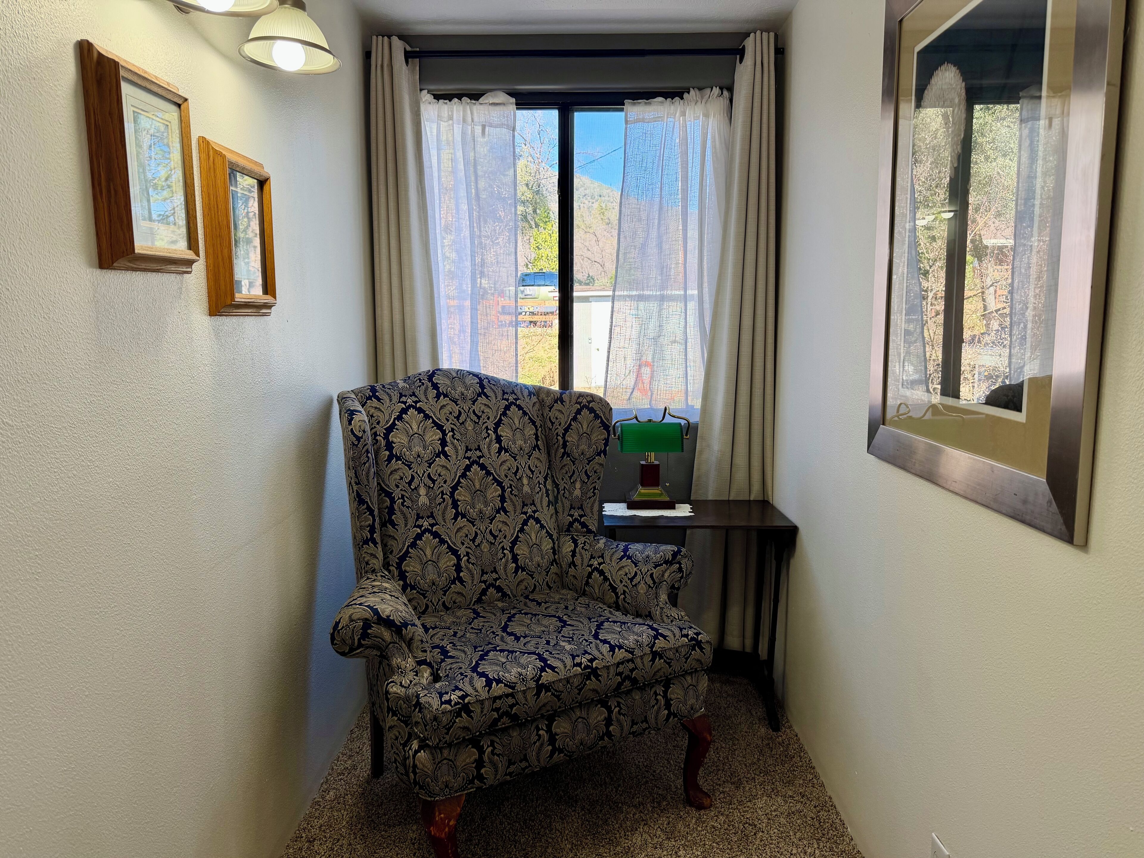 A chair with a patterned fabric, a small table with a lamp, and a window with curtains.