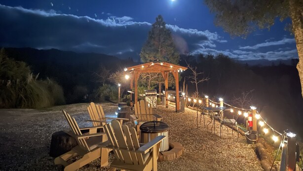 Un espace extérieur aménagé avec des chaises en bois et un foyer, situé sous une pergola illuminée de guirlandes lumineuses, le tout se détachant sur un ciel nocturne où l’on peut apercevoir la lune.