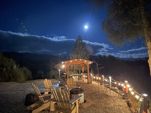 Een buitenruimte met houten stoelen en een vuurkorf, onder een pergola met lichtjes, met een zichtbare maan aan de nachthemel.