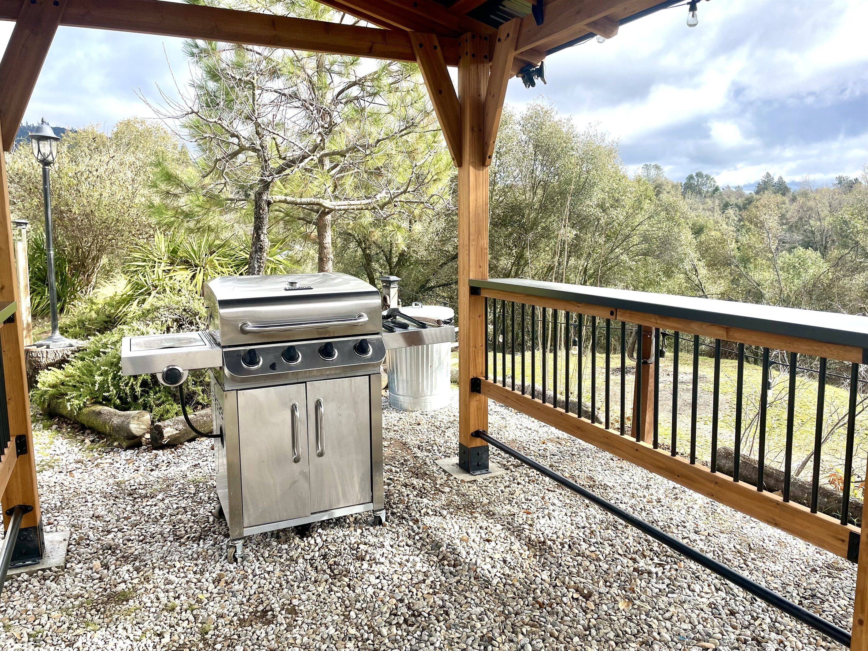An outdoor grill area with a metal grill and a stainless steel cabinet, surrounded by gravel and trees.