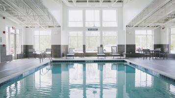 Une piscine intérieure à l’eau claire, entourée de chaises longues et de tables, avec de grandes fenêtres et un panneau indiquant « Il est interdit de plonger ».