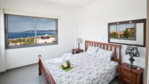 Ein Schlafzimmer mit Bett, Nachttisch, Lampe, Spiegel und Fenster mit Blick auf das Meer und die Hügel.