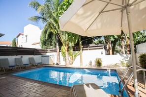 Ein Schwimmbecken mit Liegestühlen und einer überdachten Liegefläche, umgeben von Palmen und einem weißen Gebäude.