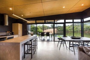 A modern kitchen with a large island, dining area with chairs, and large windows overlooking a natural landscape.