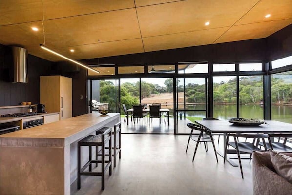 A modern kitchen with a large island, dining area with chairs, and large windows overlooking a natural landscape.