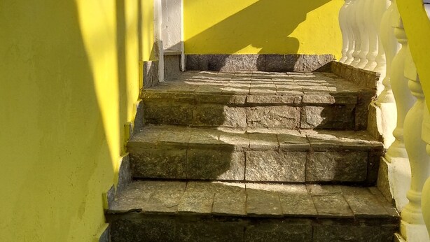 Une série de marches en pierre menant à une balustrade blanche, avec un mur jaune d’un côté et un ciel dégagé en arrière-plan.