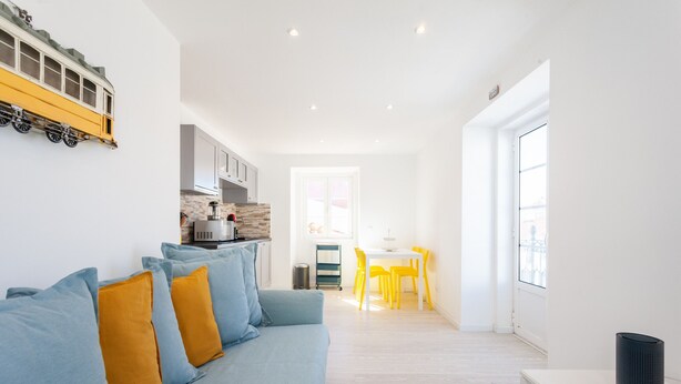 Un salon moderne avec un canapé, un coin repas avec des chaises jaunes et une cuisine équipée d’une machine à café.