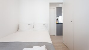 Une chambre à coucher au style minimaliste, avec un grand lit, une petite table de chevet et une vue sur un espace cuisine compact.