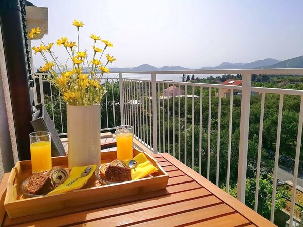 A balcony with a wooden table set with breakfast items, a vase of yellow flowers, and a scenic view of mountains and greenery.