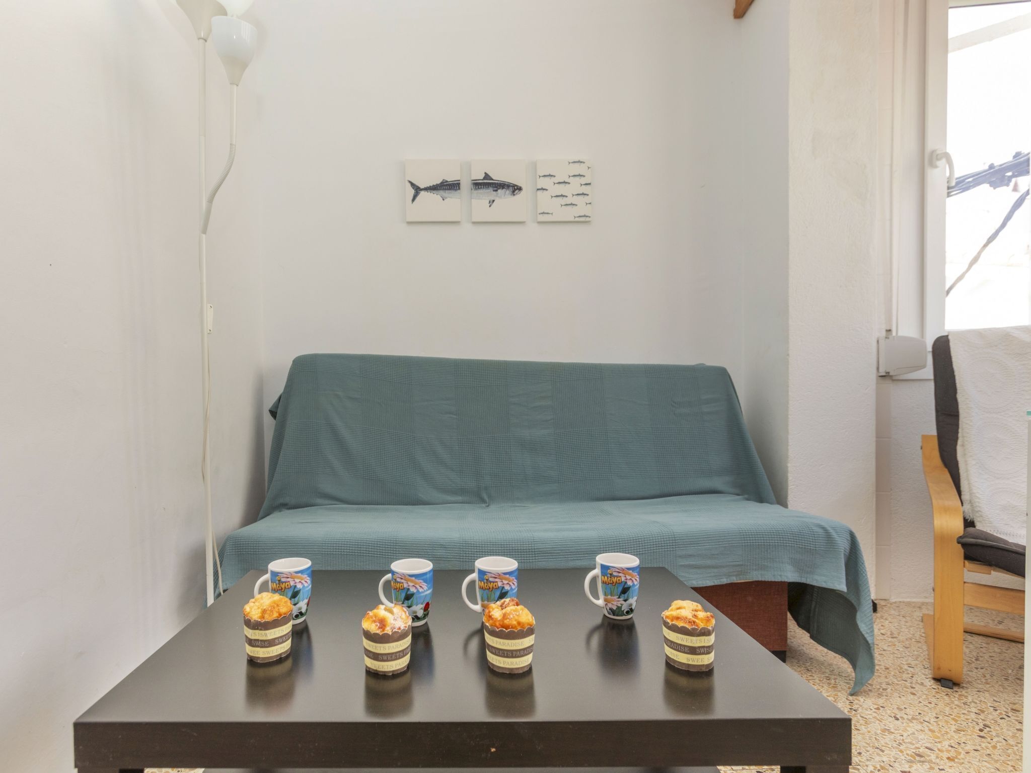A room with a teal sofa, a small table with five mugs, and three small pastries. There are two wall-mounted pictures of fish above the sofa.