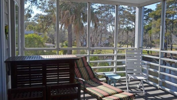 A sunroom with large windows, a wooden bench, a striped lounge chair, and a small table. - The Colony III 204 - Walk to beach in Surfside Beach (Surfside Beach)