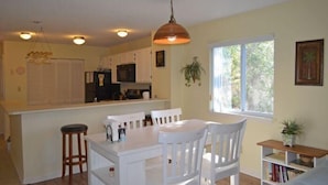 A kitchen with white cabinets, a central island with stools, a hanging pendant light, and a window with a view of greenery. - The Colony III 204 - Walk to beach in Surfside Beach (Surfside Beach)