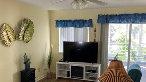 A living room with a flat-screen TV, leather sofas, a ceiling fan, decorative wall art, and a view of the outdoors through large windows. - The Colony III 204 - Walk to beach in Surfside Beach (Surfside Beach)