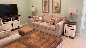 A living room with a beige sofa, wooden coffee table, and a TV on a wooden stand. There are two wall-mounted lamps and framed artwork above the sofa. - 2BR/2BA - Availability now! 
- Clean
- Close to the beach
 - Sleeps 6 (Mexico Beach)