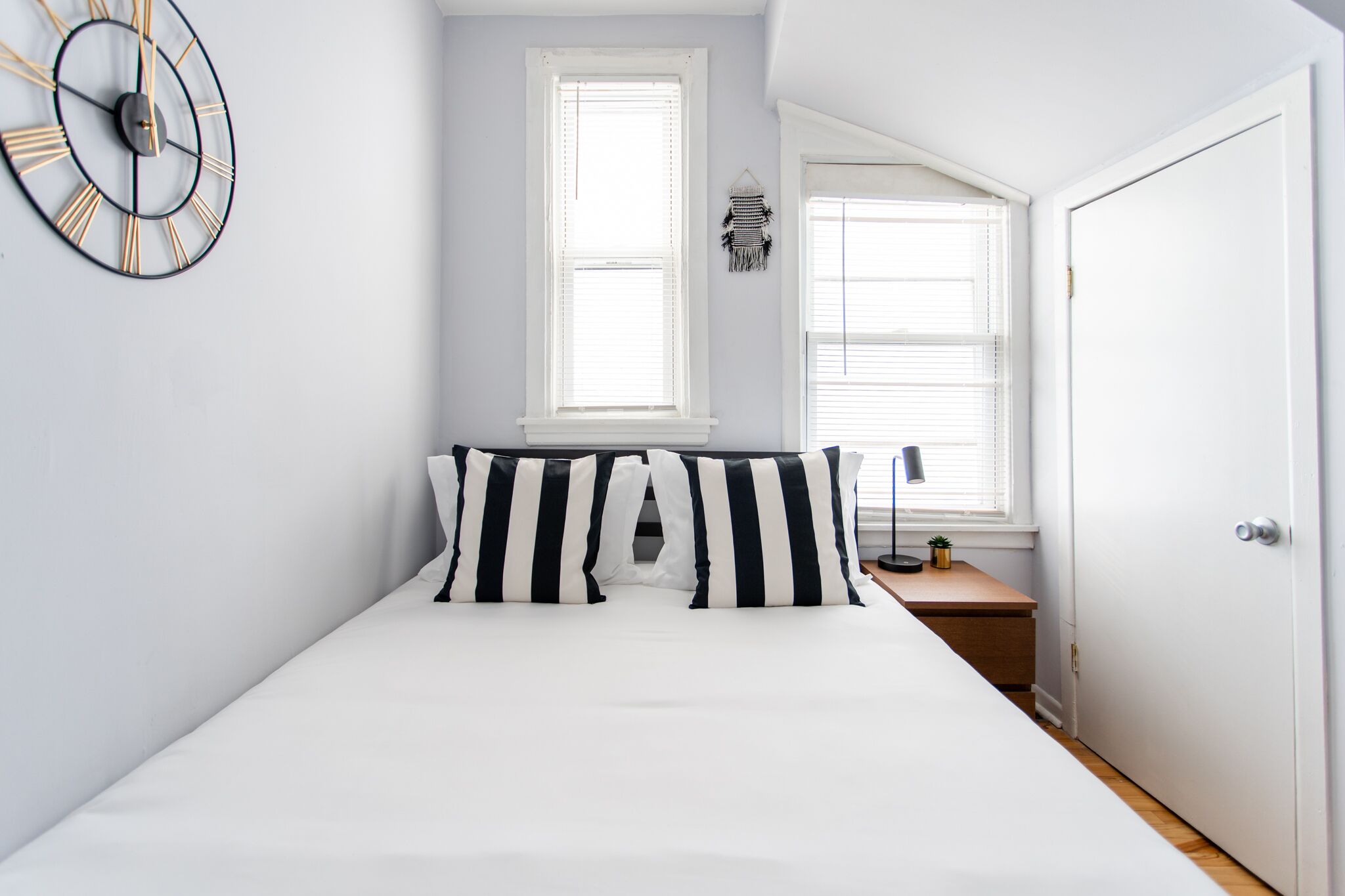 Une chambre à coucher avec un lit, deux oreillers à rayures noires et blanches, une horloge murale et deux fenêtres équipées de stores.