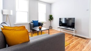 A modern living room with a grey sofa, a coffee table, a TV on a stand, and a plant. - 1R - ❤️ Low Prices! Stylish 2BR Apartment in Logan Square! (Chicago)