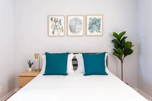 A neatly arranged bedroom with a bed, two pillows, a bedside table with a lamp, a small plant, and three framed artworks on the wall.