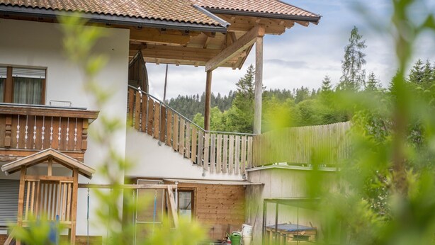 Ein zweistöckiges Haus mit Holzbalkon, einer Holzterrasse und Blick auf einen bewaldeten Bereich.