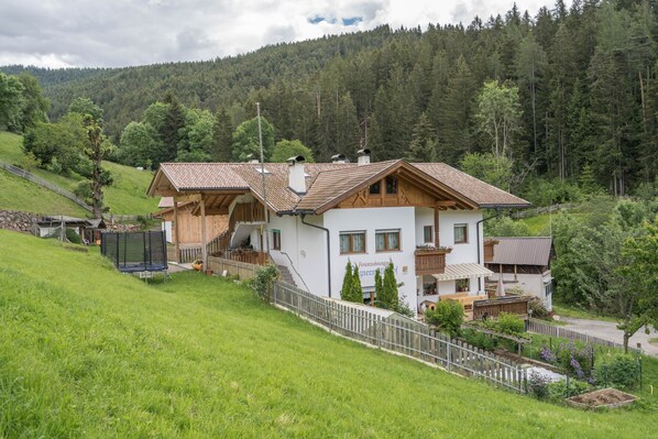 Une maison à deux étages, dotée d’un toit en pente, d’une cour clôturée et offrant une vue sur une colline boisée.