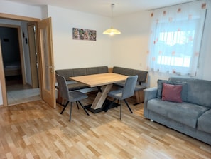 A room with a wooden dining table, grey upholstered bench seating, a grey sofa, a pendant light, and a window with curtains.