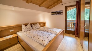 A bedroom with a wooden ceiling, a bed with floral bedding, a nightstand, a window with a view of trees, and a door leading to an outdoor area. - Holiday Apartment "Boznermüllerhof-Sonnenblume" with Balcony and View into Nature (Vöran)