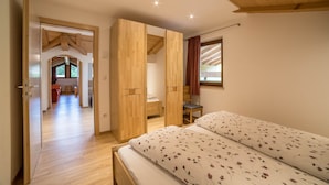 A bedroom with a wooden ceiling, a bed with floral bedding, and a view of a kitchen and dining area through an open door. - Holiday Apartment "Boznermüllerhof-Sonnenblume" with Balcony and View into Nature (Vöran)