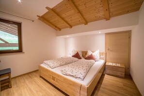 A bedroom with a wooden ceiling, a bed with floral bedding, a nightstand, a wooden bench, and a window with a view of a grassy area.