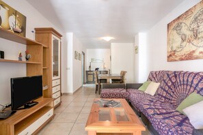 Een woonkamer met een televisie, een houten tv-meubel, een bank en een eethoek op de achtergrond.