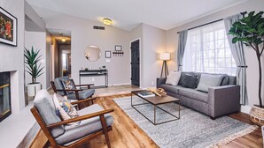 Una sala moderna con un sofá gris, una mesa de centro de madera y un tapete estampado en gris. A la izquierda hay una chimenea, a la derecha una planta grande y en la pared, un cuadro enmarcado.