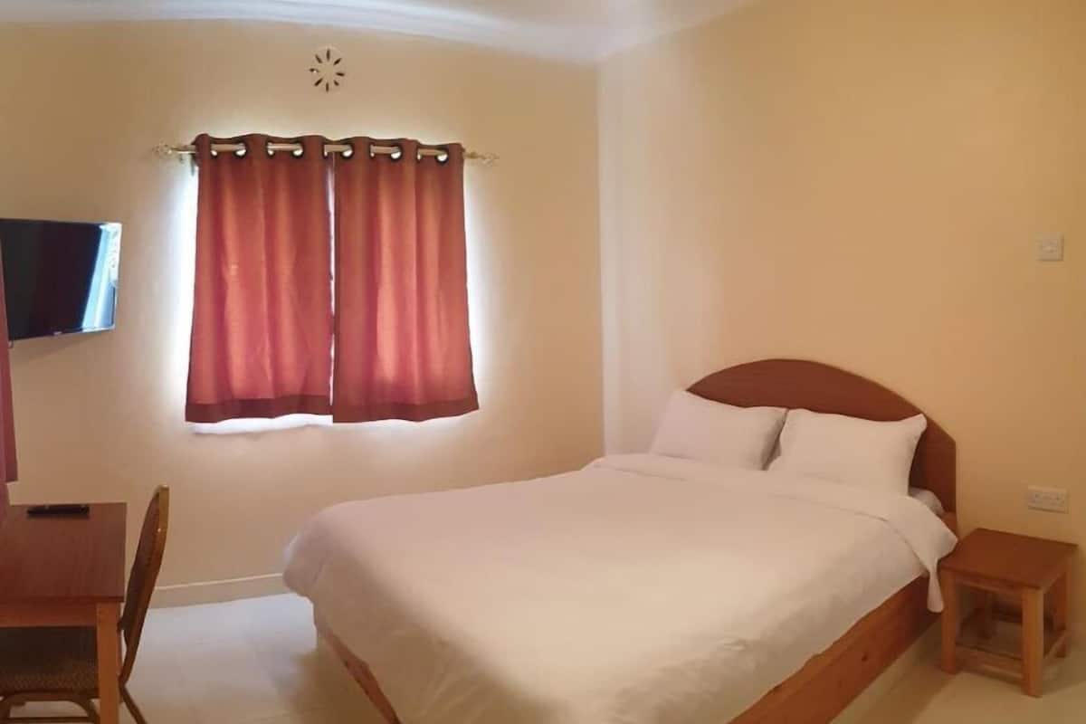 A bedroom with a bed, a wooden nightstand, a desk, a chair, and a window with curtains.