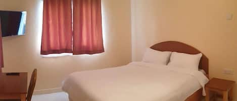 A bedroom with a bed, a wooden nightstand, a desk, a chair, and a window with curtains.