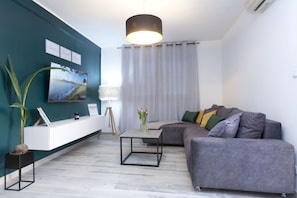 Ein modernes Wohnzimmer mit einem grauen Sofa, einem weißen Konsolentisch, einem schwarzen Couchtisch, einer Topfpflanze und einem großen Fenster mit leichtem Vorhang.